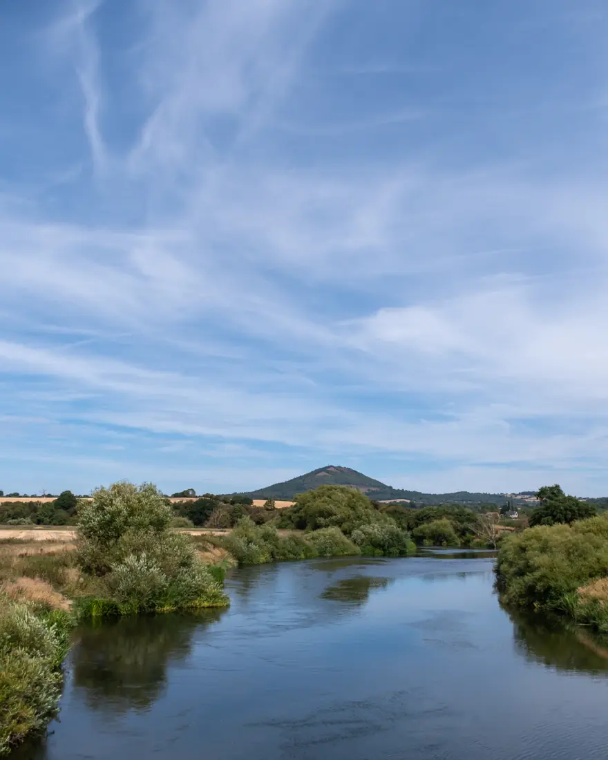 A calm river running towards a wooded hill under wispy cloud