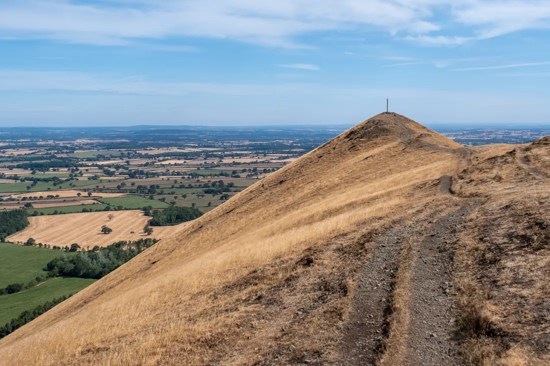 A footpath climbing a bare, sun-baked hill topped with a mast, with farmland below