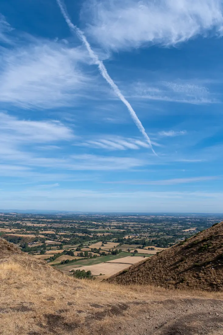 A vapour trail across a deep blue sky above patchwork farmland between two hillsides